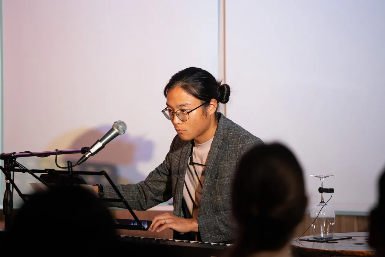 Aaron at a keyboard and laptop, one hand on the controls, with a microphone in front of him and an upturned wine glass on a table beside him.