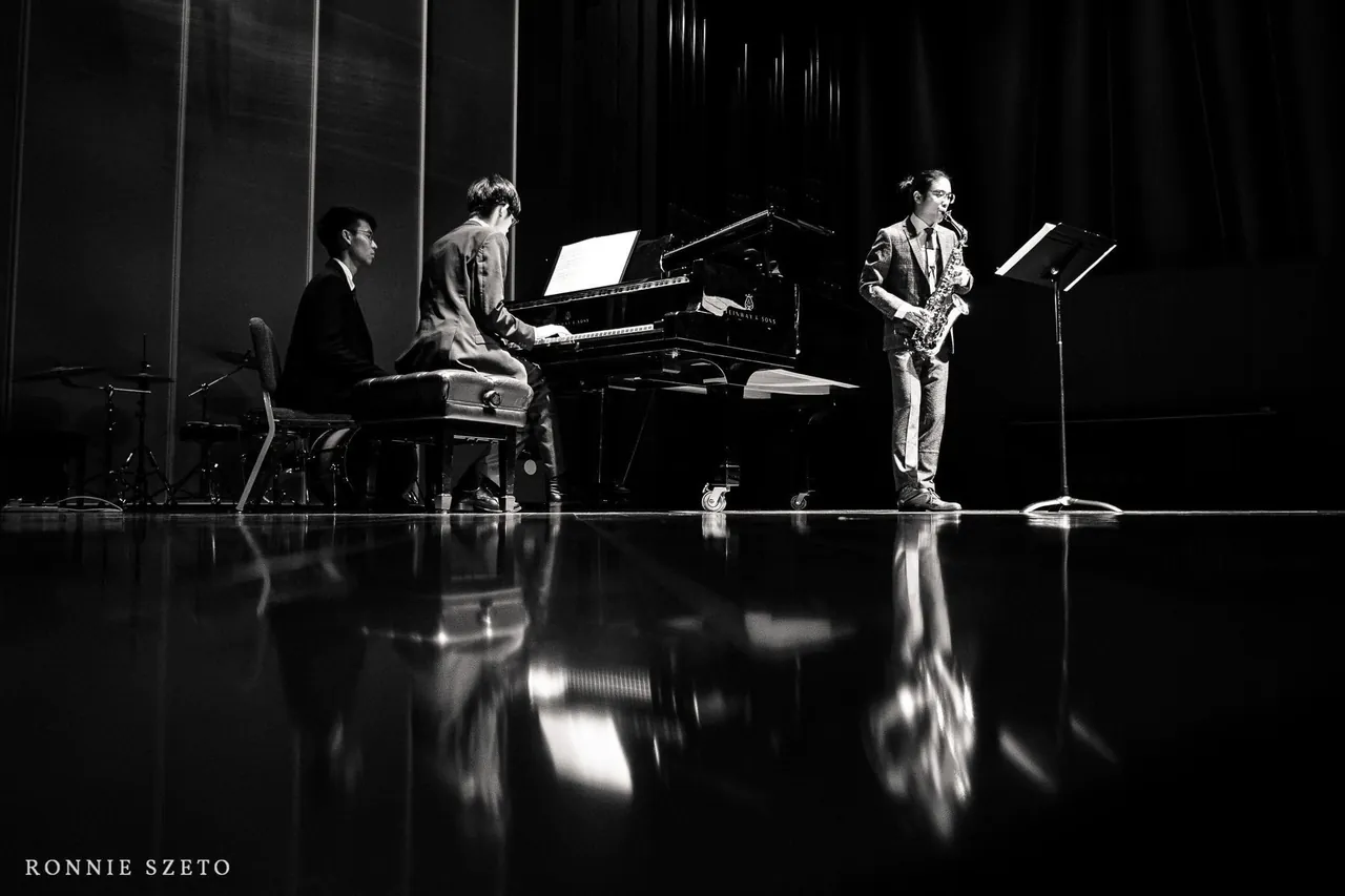 Aaron on alto saxophone beside a grand piano with two musicians at it and a drum kit behind, their reflections on the polished stage floor.