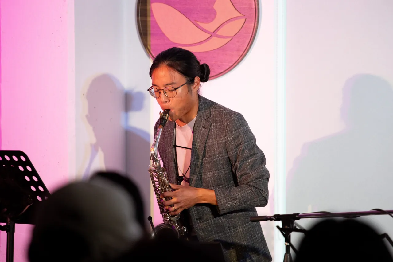Aaron playing alto saxophone with his eyes closed, in front of a wooden bird emblem with pink light on the wall.