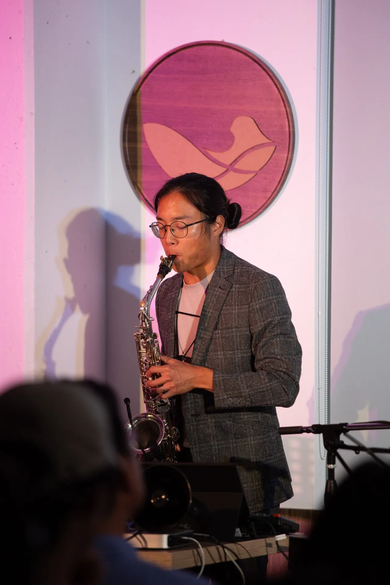 Aaron playing alto saxophone in front of a round wooden bird emblem, lit in soft pink.